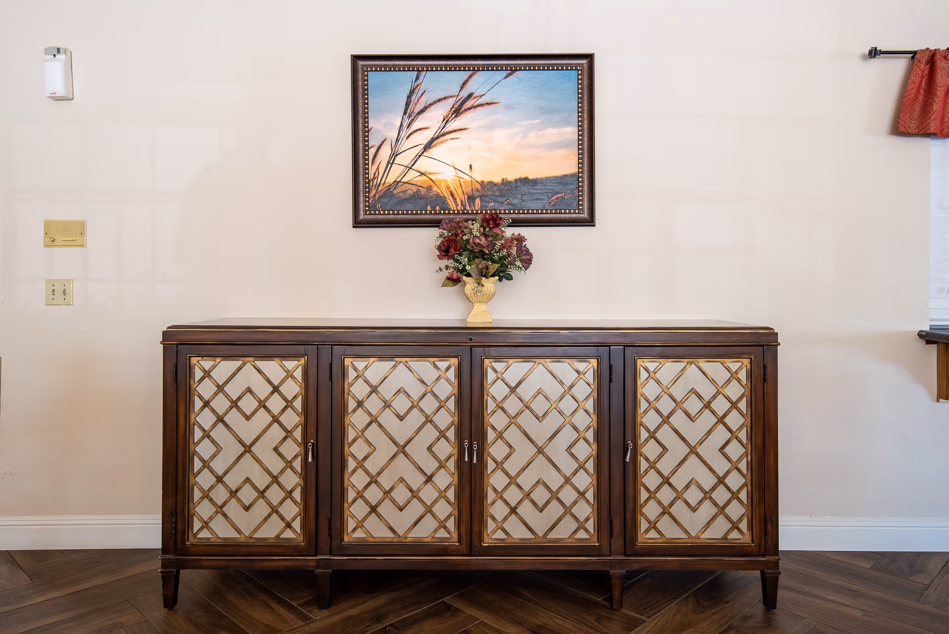 Ornate wooden sideboard with a vase of flowers beneath a framed sunset print on a neutral wall.