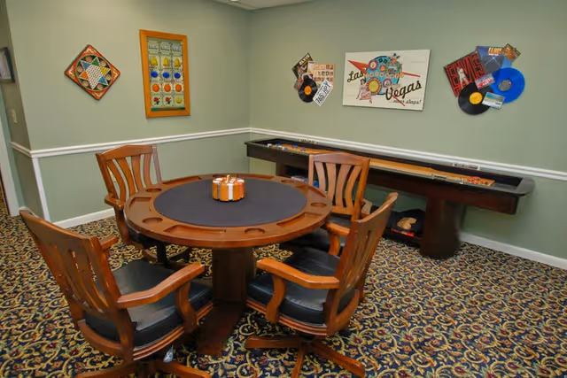 A game room with a round wooden table surrounded by four wooden chairs with black cushions. The table has cup holders and a small decorative item in the center. Behind the table is a shuffleboard table against a light green wall decorated with colorful framed artwork and vinyl record wall hangings. The floor is covered with a patterned carpet.