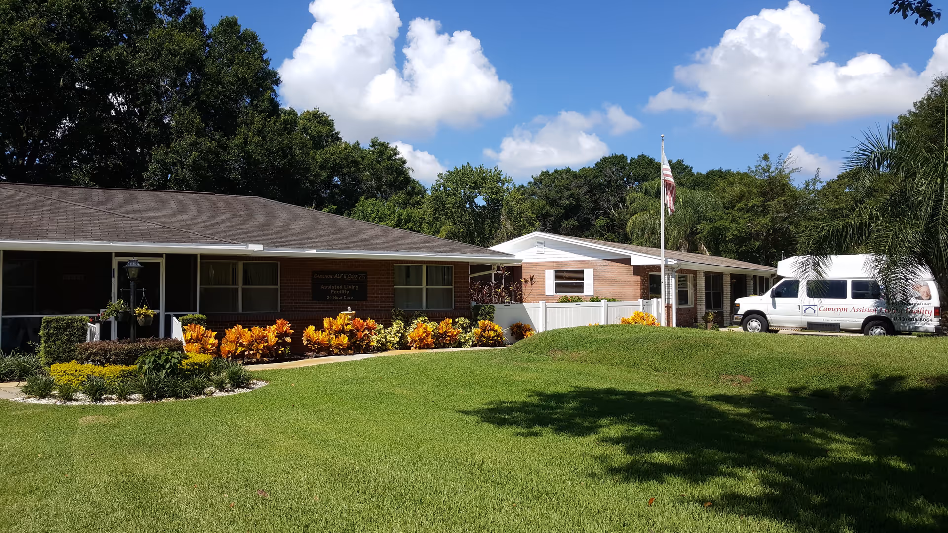 Exterior view of Cameron Assisted Living Facility showing a single-story brick building with a well-maintained lawn, colorful flower beds, an American flag on a flagpole, and a white van parked near the entrance under a blue sky with scattered clouds.
