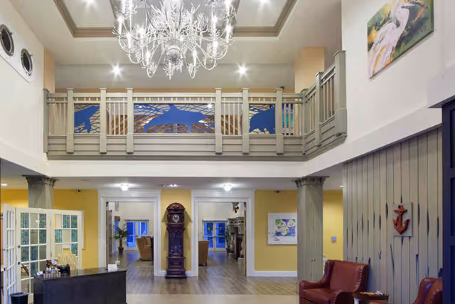 Interior view of a senior living facility lobby with a high ceiling, a large chandelier, a balcony with decorative railings featuring dolphin designs, a grandfather clock, and seating areas with red leather chairs. The walls are painted yellow and white, and there is artwork on the walls.