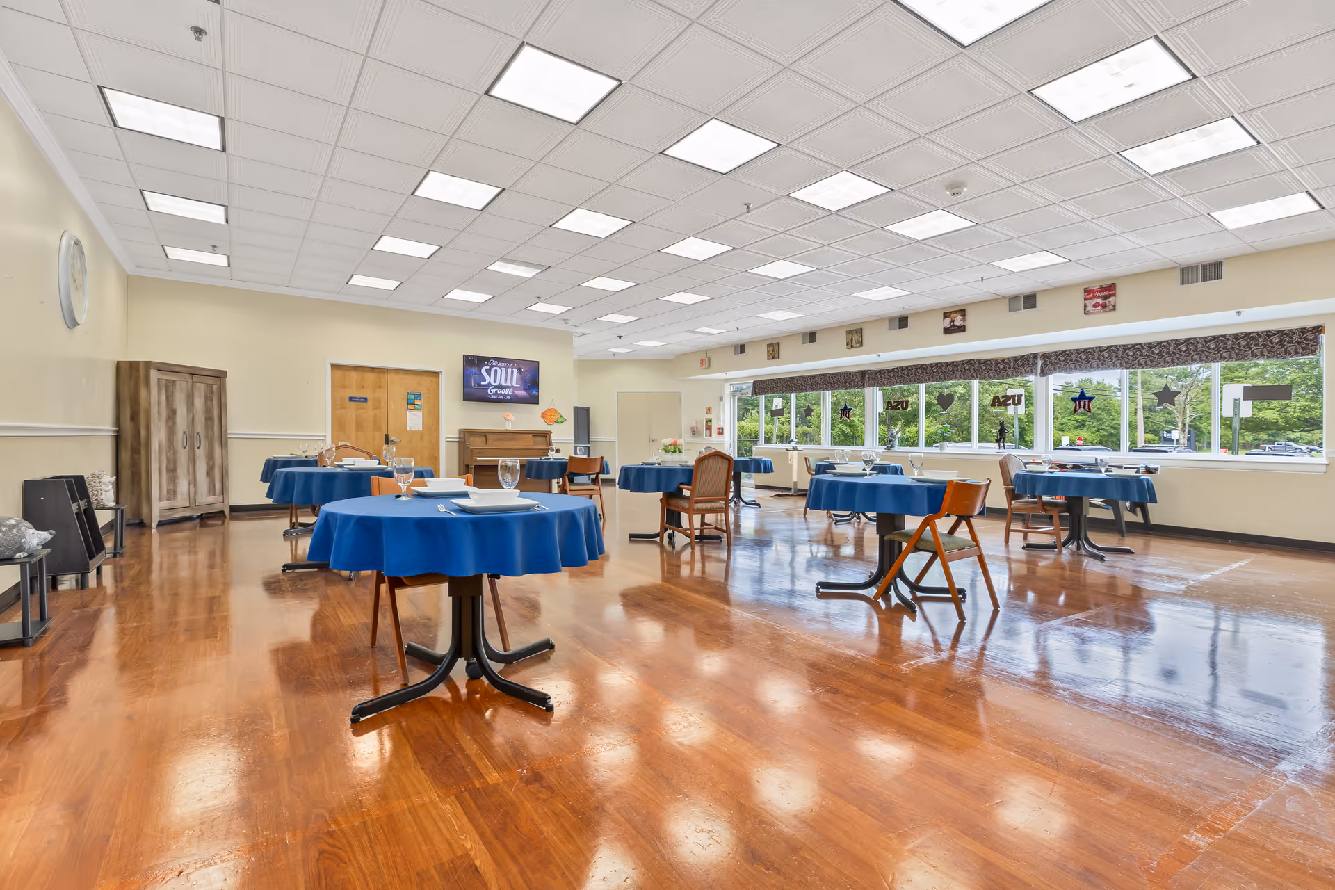 A spacious dining room with several round tables covered with blue tablecloths, each set with plates, glasses, and silverware. The room has large windows along one wall letting in natural light, wooden floors, and a ceiling with multiple fluorescent lights. There is a wooden cabinet and a piano against one wall, and decorations on the windows and walls.