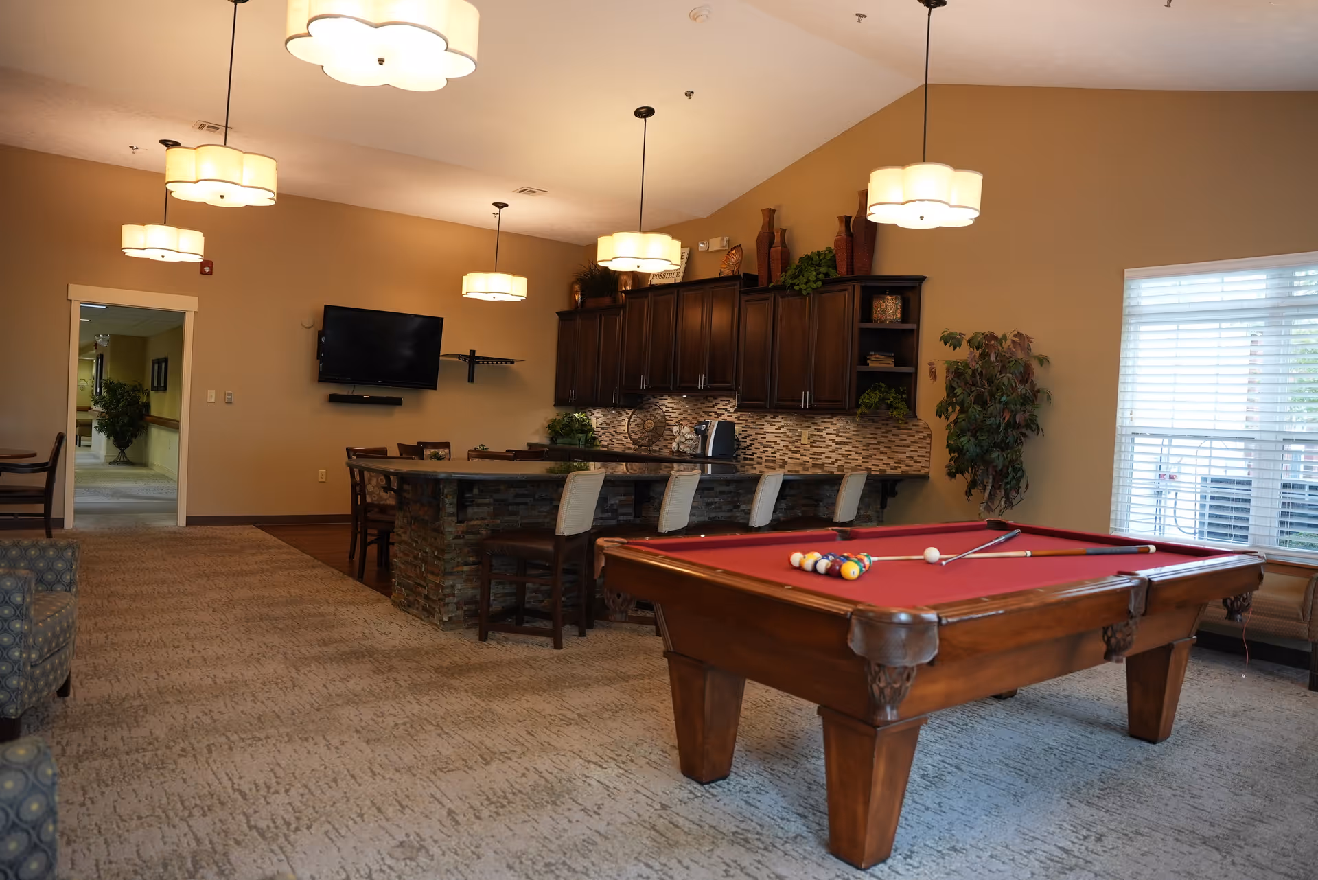 Community lounge with a red pool table in the foreground, bar seating and a kitchenette under pendant lights.
