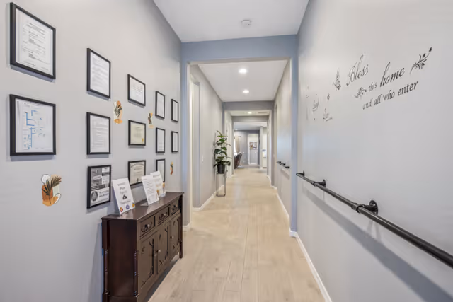 A well-lit hallway in an assisted living facility with light wood flooring and light gray walls. On the left wall, there are framed certificates and documents above a dark wooden cabinet. The right wall features a handrail and decorative wall text that reads 'Bless this home and all who enter.' The hallway extends into the distance with additional rooms and plants visible.