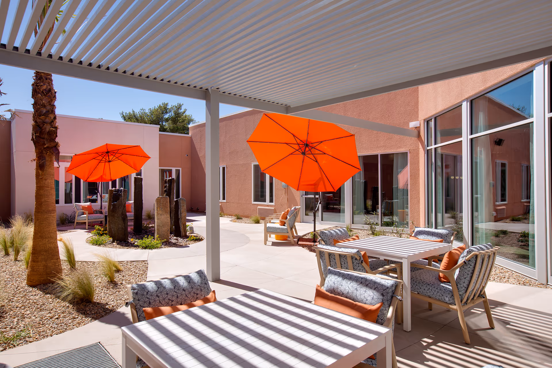 Outdoor patio area at Lumina Las Vegas featuring white tables and cushioned chairs with orange pillows under a white pergola. Bright orange umbrellas provide shade near a small garden with tall stone sculptures and desert landscaping including palm trees and gravel.