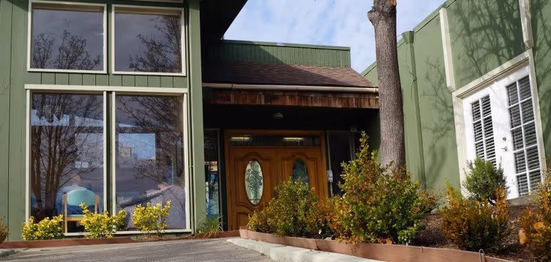 Entrance to a building with green exterior walls, large windows reflecting trees, a wooden double door, and landscaped bushes along the walkway.