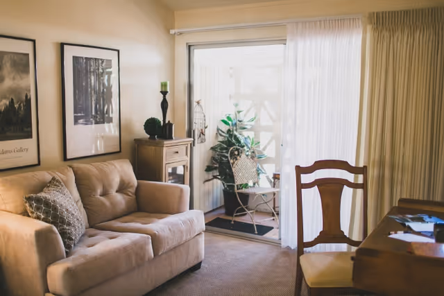 A cozy living room with a beige sofa adorned with a patterned pillow, two framed pictures on the wall, a small wooden cabinet with decorative items including a green candle and a plant, and a sliding glass door leading to a small outdoor patio with a metal chair and plants. A wooden chair and desk with papers are partially visible on the right side.