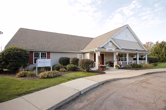 Exterior view of a single-story building with a gable roof, white siding, and a covered front porch with columns. There are shrubs and flowers planted along the front, a sidewalk leading to the entrance, and a sign that reads 'Brookdale Monroe'. The sky is partly cloudy.