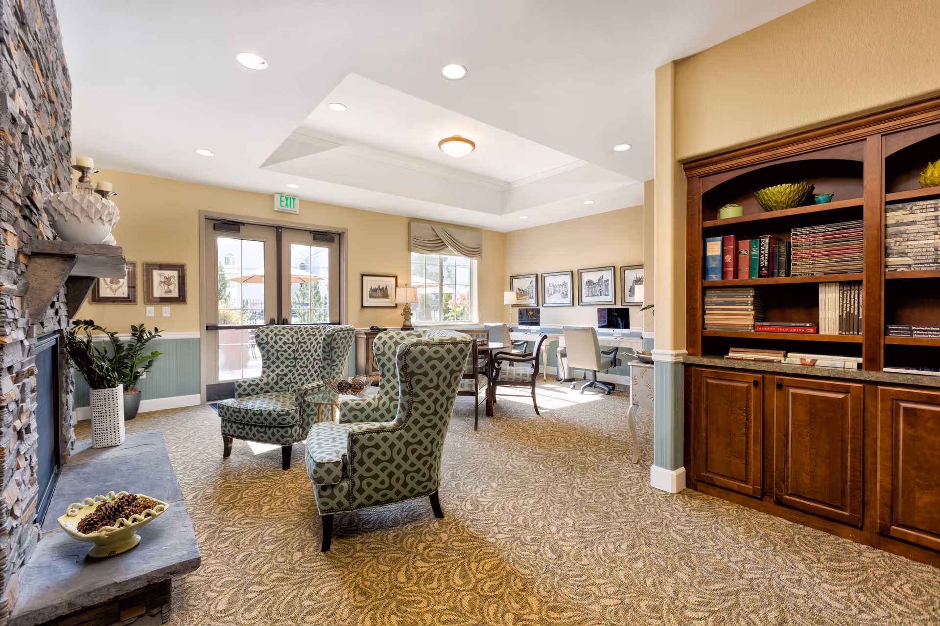 A bright and cozy living room area in an assisted living facility featuring two patterned armchairs around a small glass table, a stone fireplace with decorative items on the mantel, a large wooden bookshelf filled with books and decorative pieces, and a workspace with computers and chairs near windows letting in natural light.