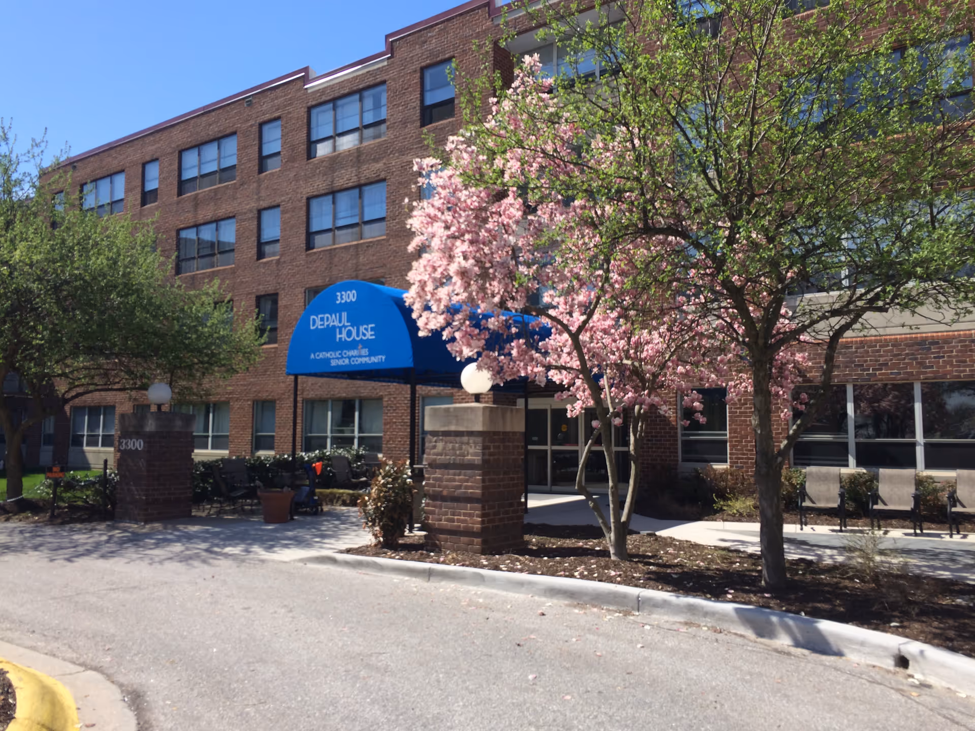 Front entrance of a brick senior living building with a blue 'DePaul House' canopy and flowering trees.