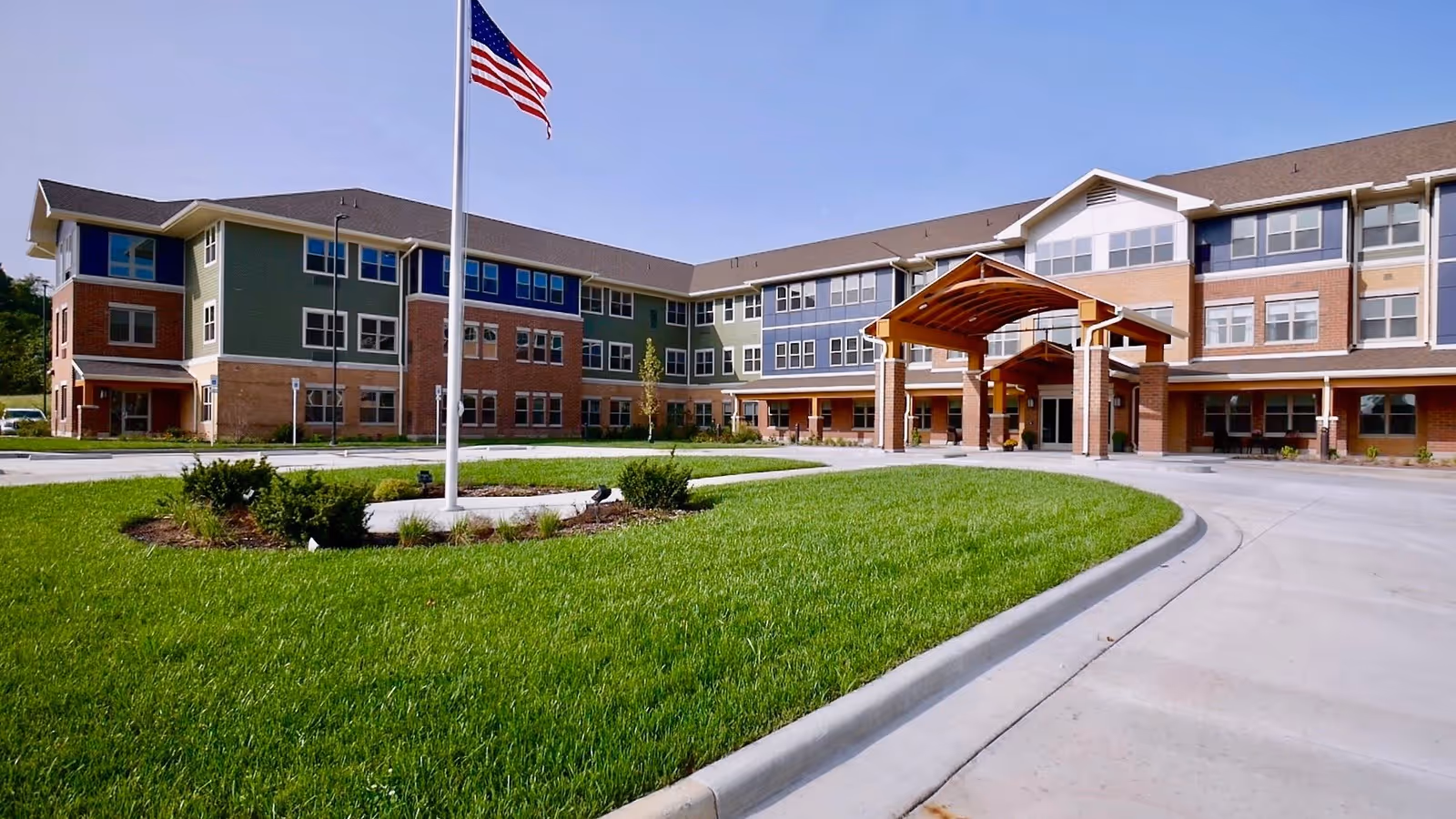 Exterior view of a large three-story senior living facility building with a covered entrance, an American flag on a flagpole, and well-maintained green lawn and landscaping under a clear blue sky.