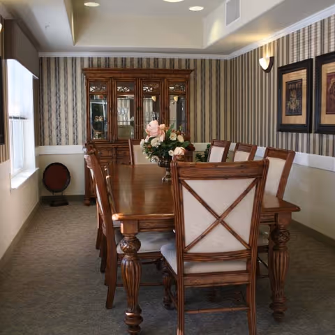 A formal dining room with a long wooden table surrounded by eight upholstered wooden chairs. A floral centerpiece is placed on the table. The room has striped wallpaper, a wooden china cabinet with glass doors displaying glassware, two framed artworks on the wall, and a window letting in natural light.