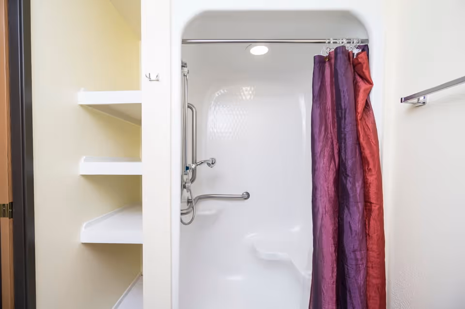 A bathroom shower area with a white built-in seat and grab bars, partially covered by a purple and red shower curtain. To the left, there are white built-in shelves, and to the right, a towel rack is mounted on the wall.