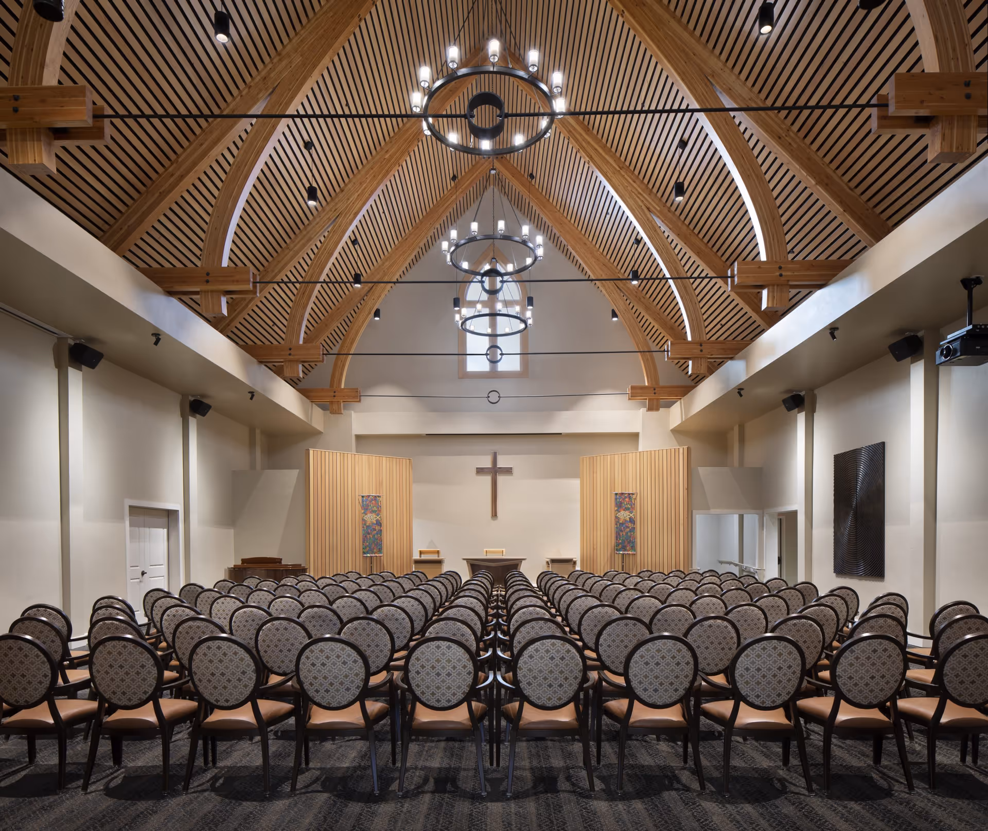 Interior view of a chapel or worship room with rows of patterned chairs facing a wooden altar with a cross on the wall. The ceiling features wooden beams and modern circular chandeliers.