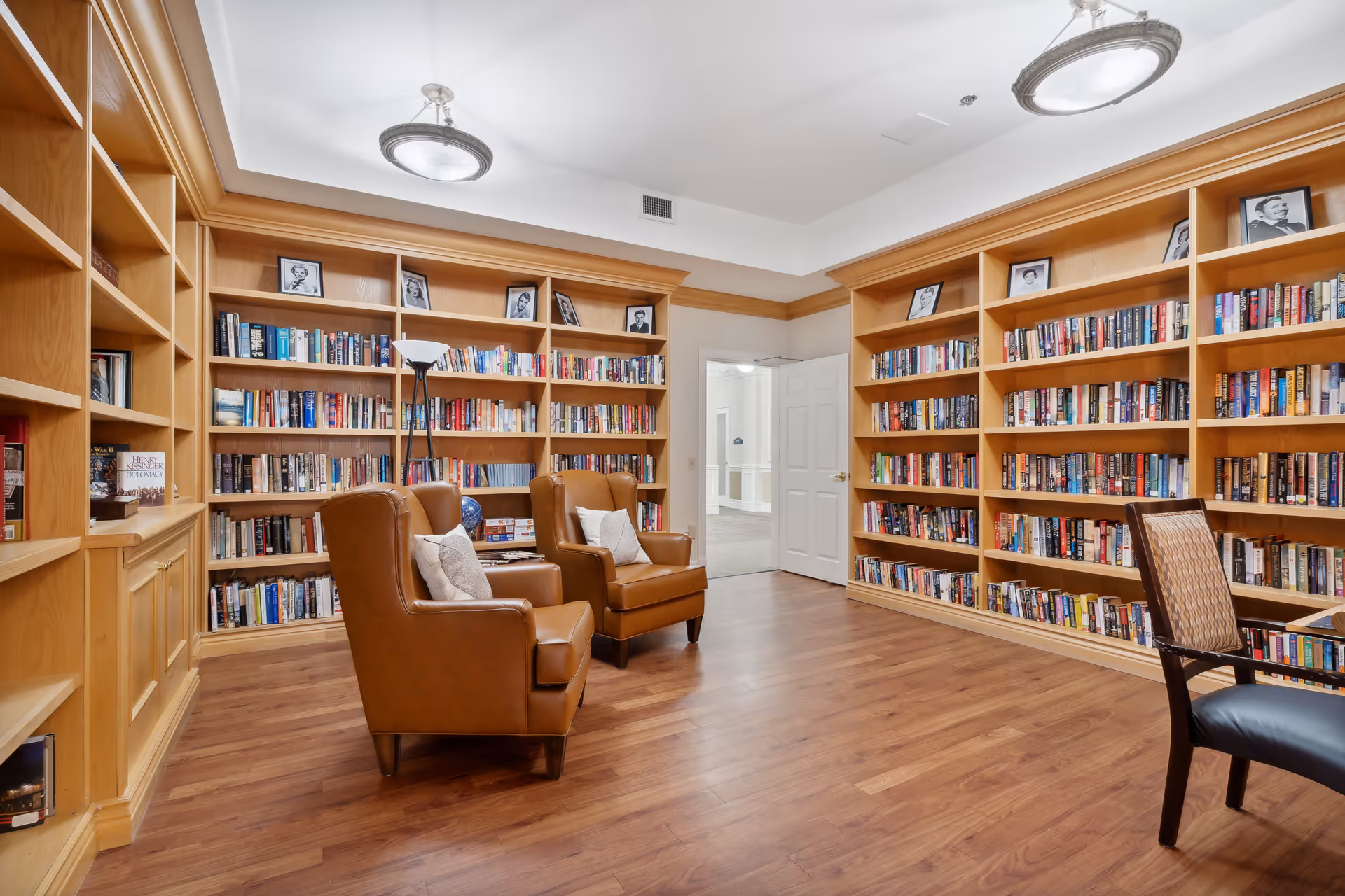 A cozy library room with wooden bookshelves filled with books lining the walls. Two brown leather armchairs with pillows are positioned in the center of the room on a wooden floor. A standing lamp is placed near the chairs, and a wooden chair with a woven backrest is partially visible on the right. The room has a white ceiling with two circular light fixtures and an open door leading to a hallway.