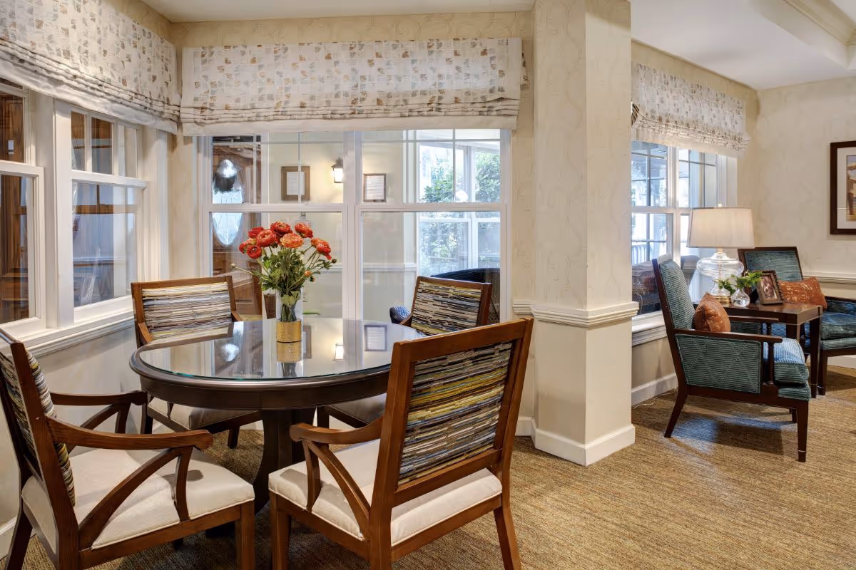 A cozy interior seating area with a round glass-top table surrounded by four wooden chairs with patterned cushions. A vase with red flowers is placed on the table. In the background, there are two armchairs with blue upholstery and orange pillows, a side table with a lamp, and large windows with patterned valances letting in natural light.