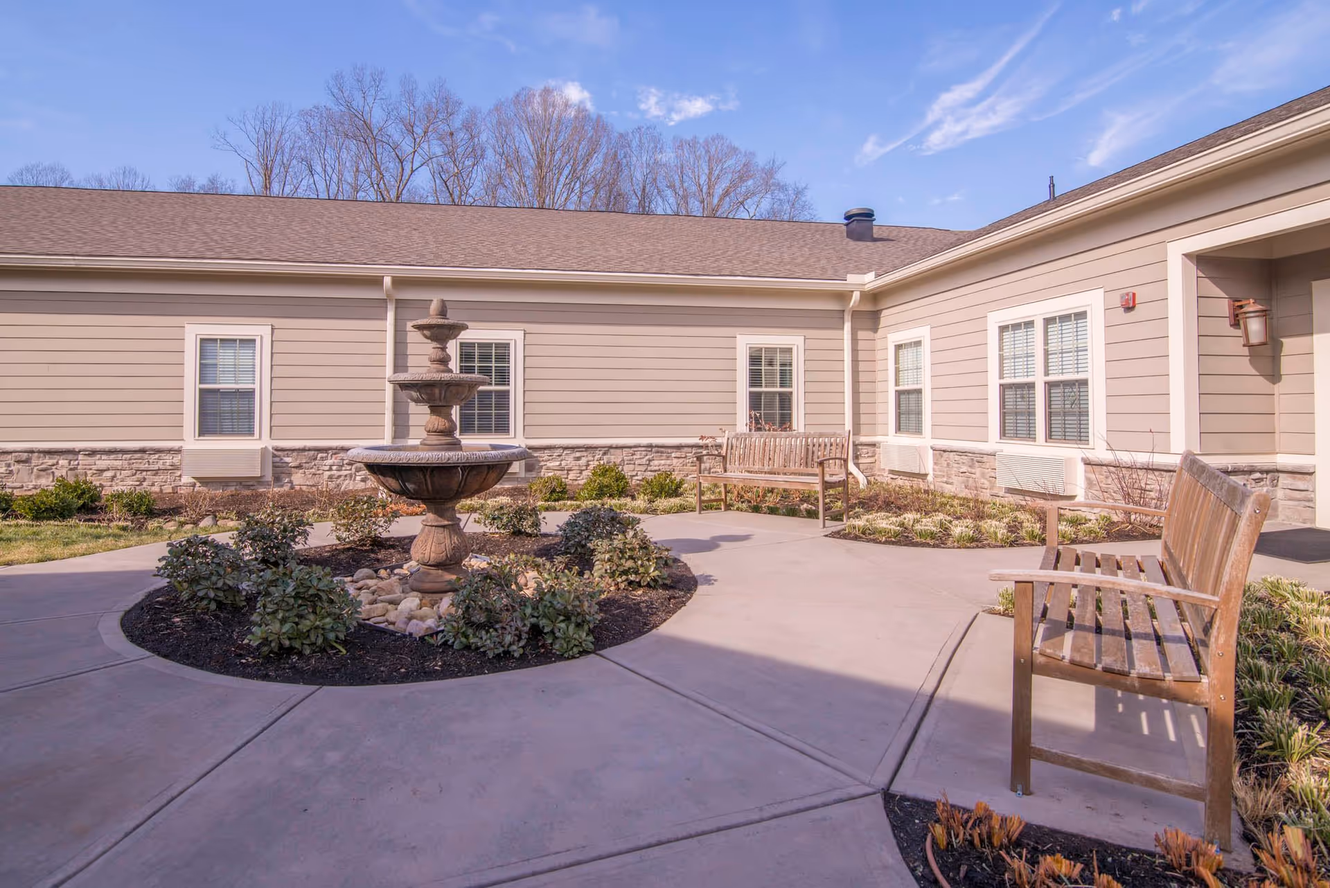 Outdoor courtyard area at Morning Pointe of Powell featuring a central tiered stone fountain surrounded by small bushes and plants, with wooden benches placed along the concrete walkway. The building exterior has beige siding with stone accents and multiple windows, under a clear blue sky.