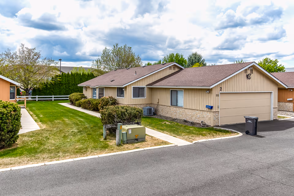 Single-story beige ranch-style building with an attached garage, green lawn, shrubs, and a paved road under a cloudy sky.