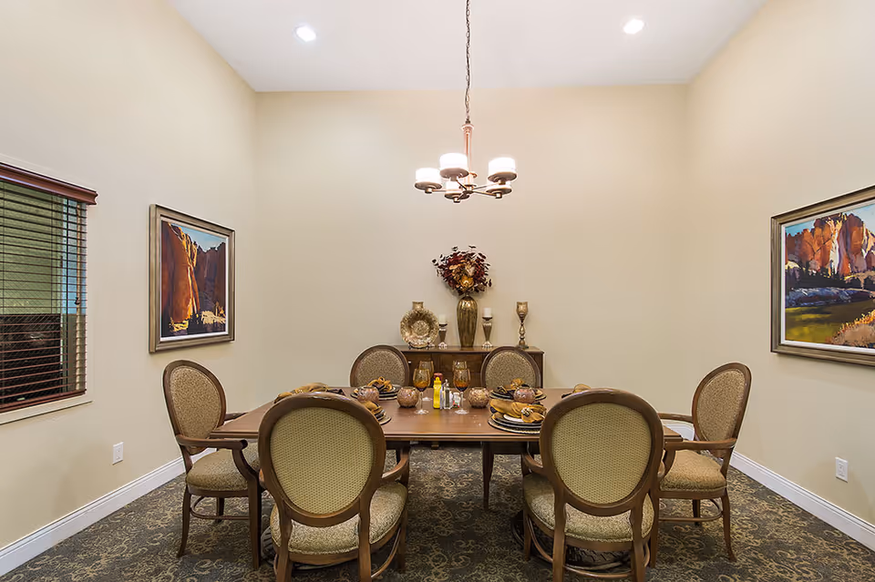 A formal dining room with a wooden table set for six people. The table has plates, glasses, and decorative items. Six upholstered chairs surround the table. The room has beige walls with two framed landscape paintings, a window with blinds on the left, a chandelier hanging from the ceiling, and a sideboard with decorative vases and candles against the back wall.