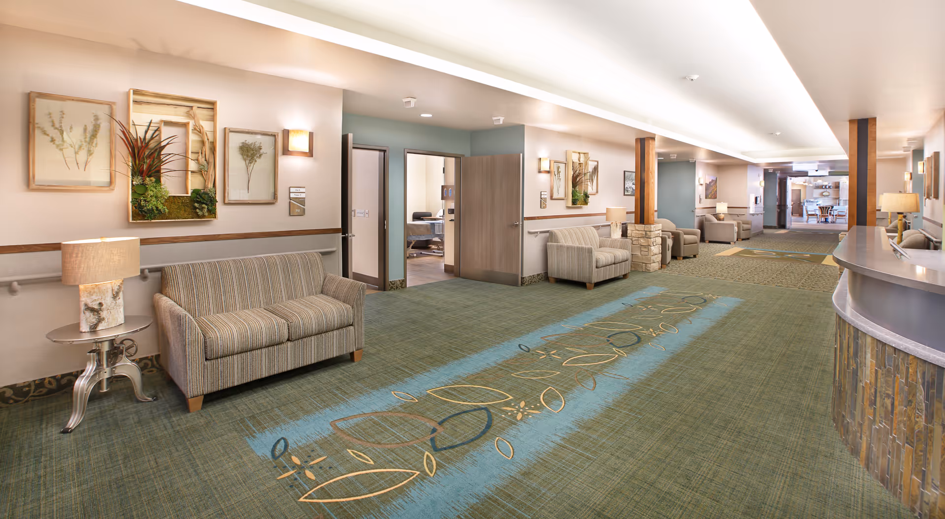 A spacious and well-lit common area in a senior living facility featuring multiple seating arrangements with armchairs and sofas along the walls. The carpet has a decorative pattern, and the walls are adorned with framed botanical artwork and wall-mounted lamps. There is a reception desk on the right side and open doorways leading to other rooms.