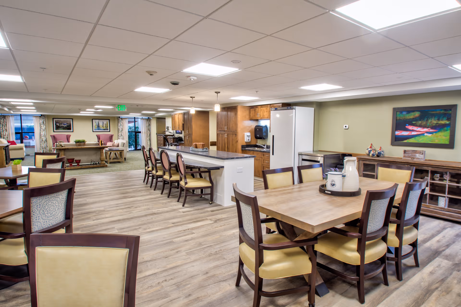 A spacious senior living facility common area featuring a dining space with wooden tables and chairs, a kitchen area with a large island and bar stools, and a lounge area with armchairs and a coffee table. The room has wood flooring, a drop ceiling with recessed lighting, and large windows with curtains allowing natural light.