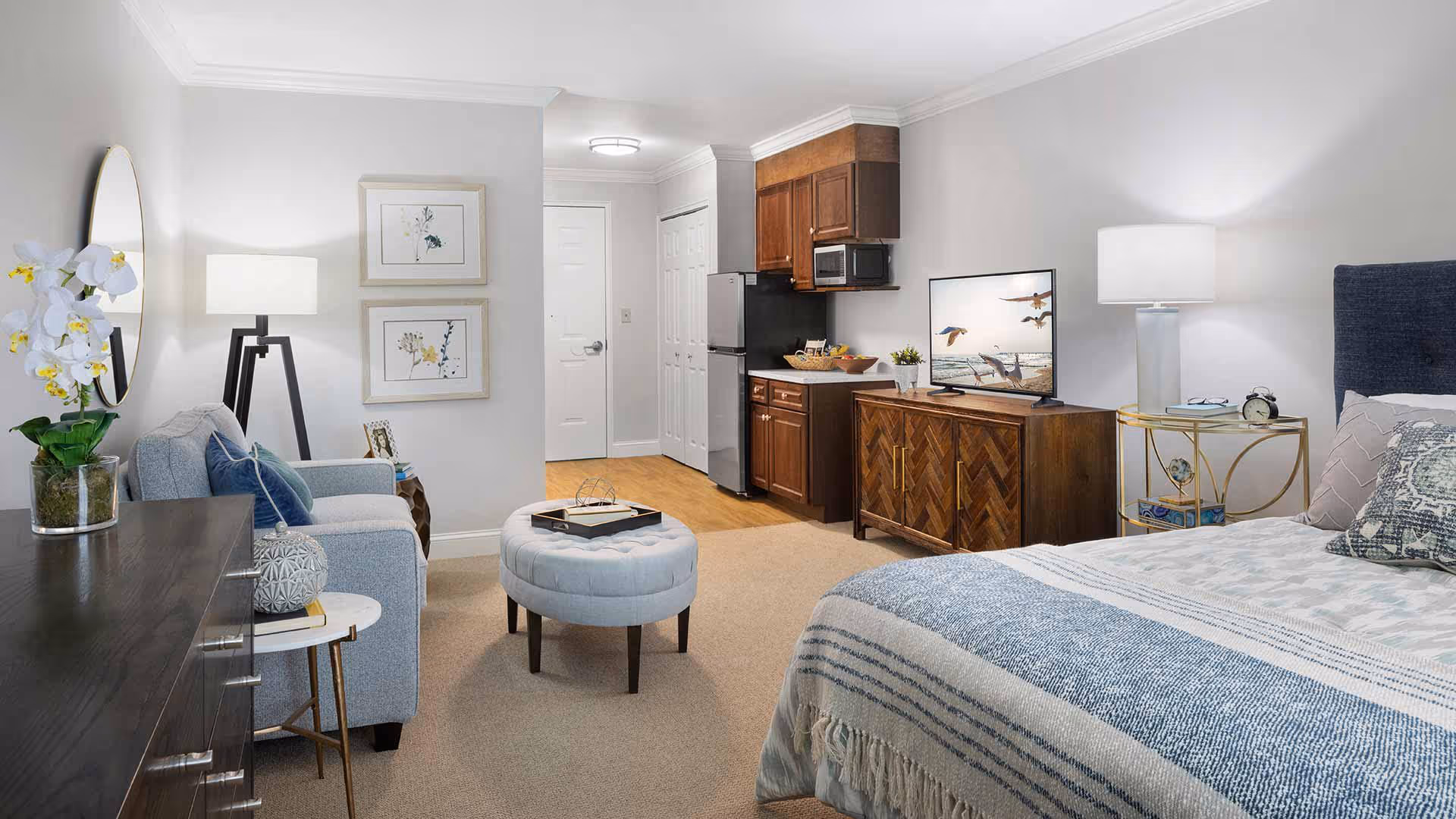 A well-lit studio apartment featuring a bed with blue and white bedding on the right, a wooden dresser with a flat-screen TV displaying flying birds, a small kitchenette with wooden cabinets, a refrigerator, and a microwave in the background. On the left, there is a light blue sofa with cushions, a round ottoman in front of it, a floor lamp, and a side table with a decorative item. The walls are painted light gray, and there are two framed floral artworks hanging near the entrance door.