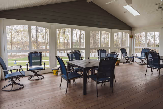 Enclosed screened porch with multiple chairs and a table overlooking a yard and pond.