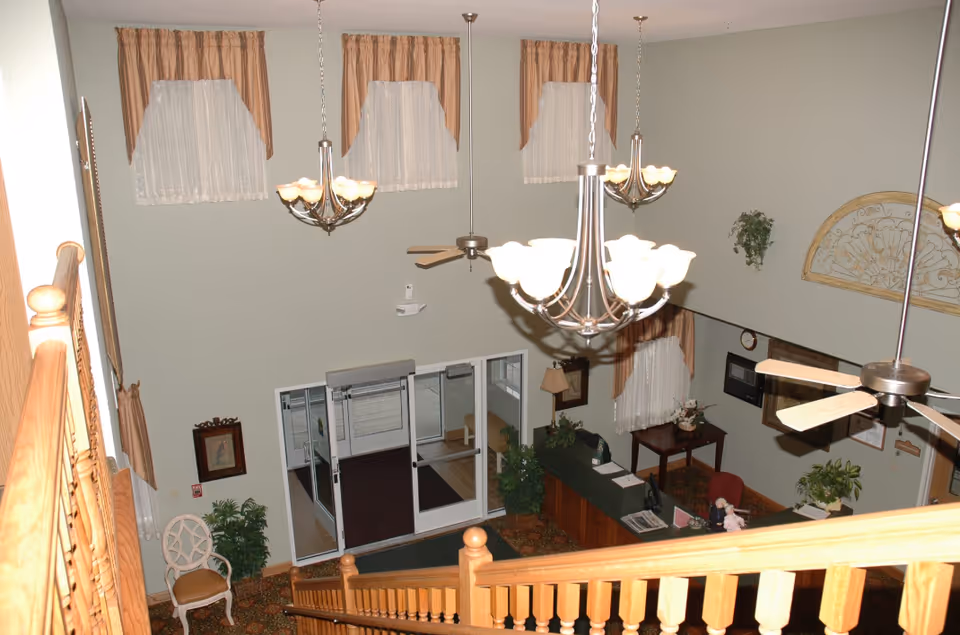 View from the top of a wooden staircase looking down into a lobby area with a reception desk, ceiling fans, chandeliers, decorative plants, framed pictures, and windows with curtains.