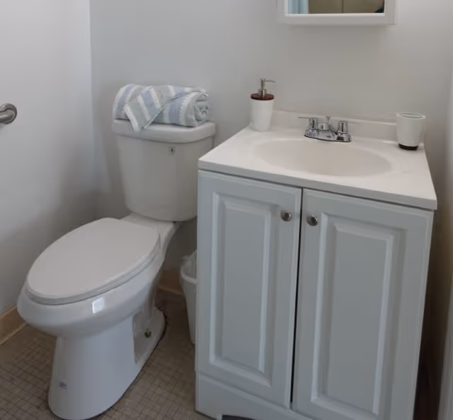 A small bathroom with a white toilet topped with a folded towel and a washcloth, next to a white vanity with a sink, faucet, soap dispenser, and a cup. A mirror is partially visible above the sink.