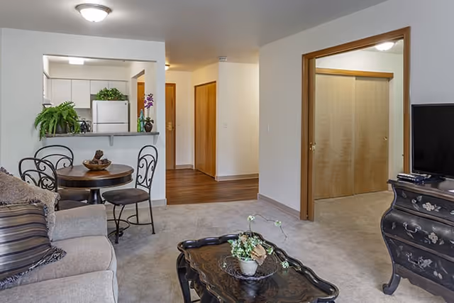 Interior view of a senior living facility apartment showing a living room with a beige sofa, a decorative coffee table with a plant, a round dining table with four chairs, and a kitchen area with white cabinets and a refrigerator. There is a hallway with wooden doors and an open doorway leading to another room.