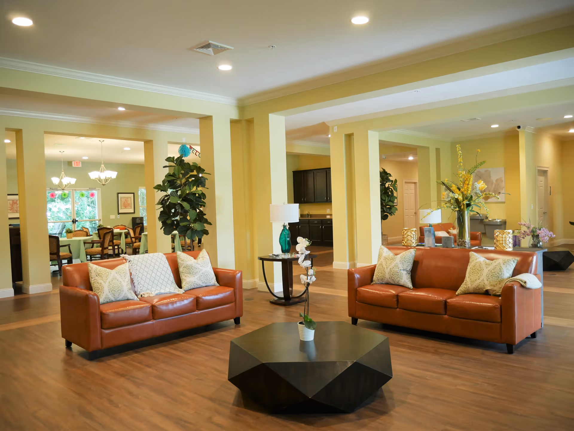 A spacious and well-lit living area in a senior living facility featuring two brown leather sofas with decorative pillows, a modern black geometric coffee table with a small potted plant, and a side table with a green glass lamp and white orchid. The room has light yellow walls, wooden flooring, and is decorated with large plants and floral arrangements. In the background, a dining area with tables and chairs is visible.