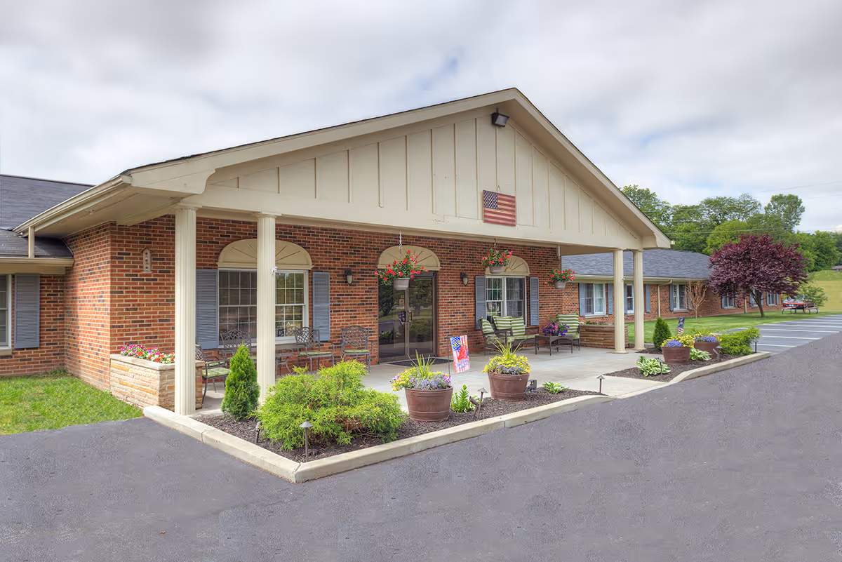 Exterior view of a single-story brick building with a covered entrance supported by white columns. There are hanging flower pots and potted plants near the entrance, along with outdoor seating. An American flag is mounted above the entrance. The surrounding area includes a parking lot and green trees in the background under a cloudy sky.