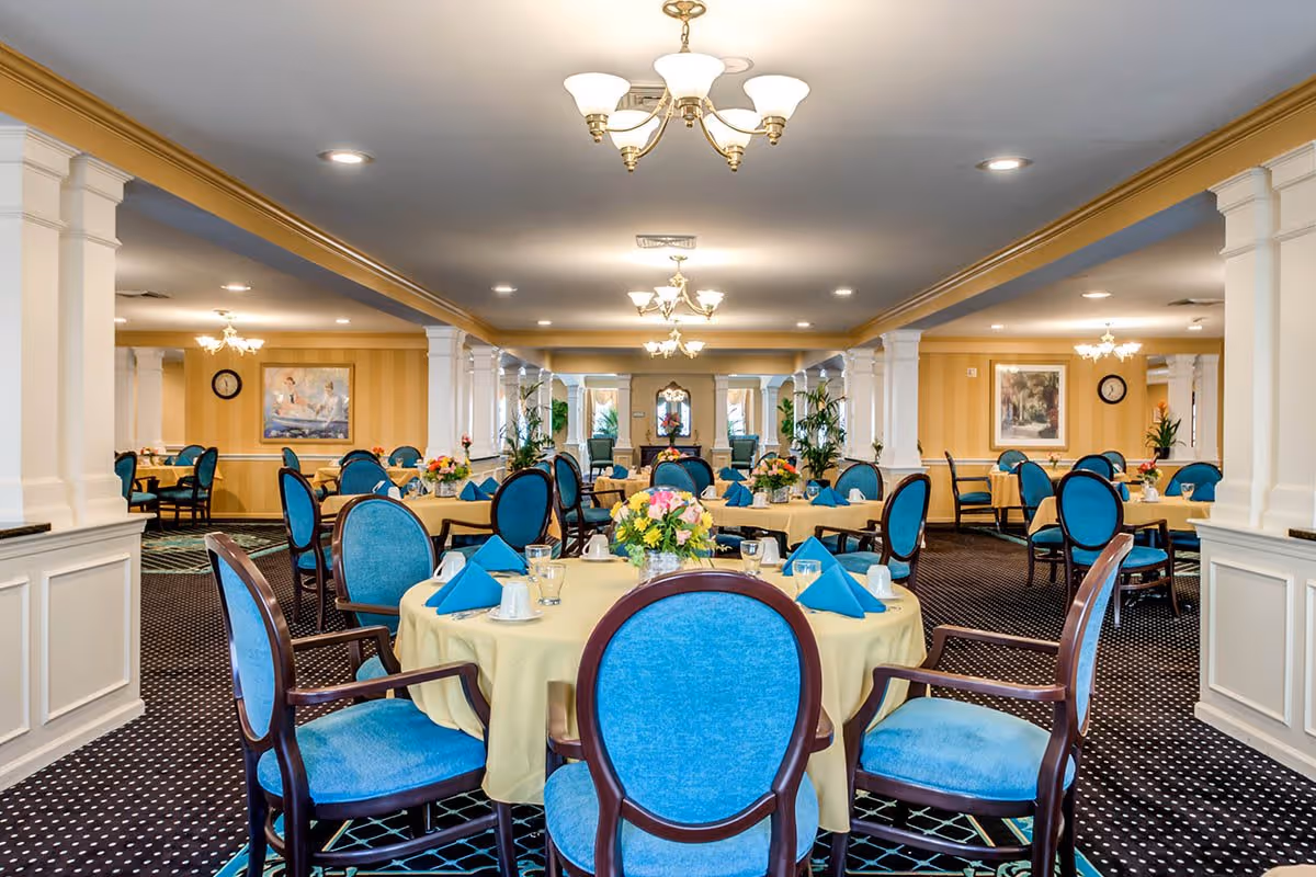 A spacious dining room with multiple round tables covered with yellow tablecloths and set with blue napkins, cups, and glasses. The chairs have blue upholstery and wooden frames. The room features white columns, chandeliers, framed artwork on the walls, and floral centerpieces on each table.