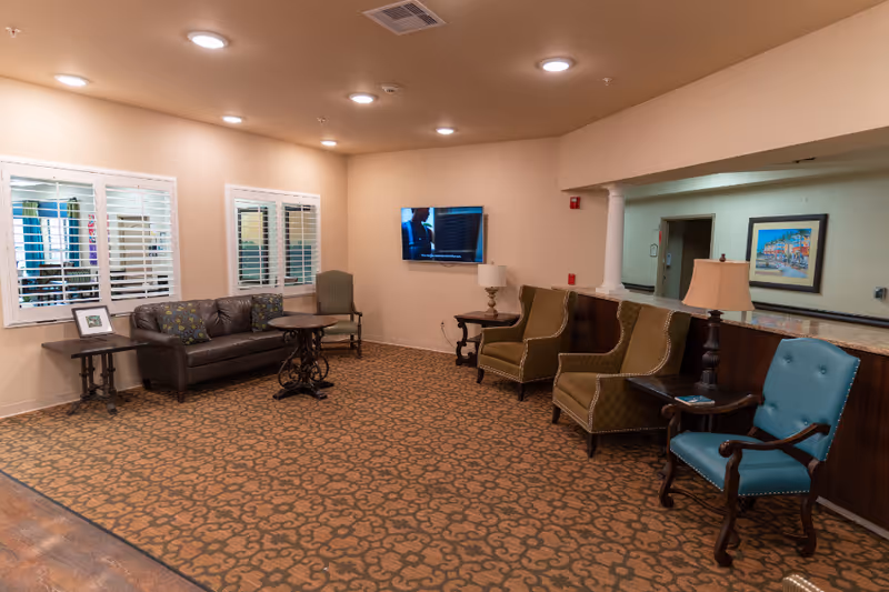 A well-lit senior living lounge with a sofa, several armchairs, side tables, a wall-mounted TV, and carpeted floor.