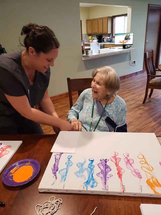 A caregiver and an elderly woman are seated at a table in a room with wooden flooring and a kitchen area in the background. The elderly woman is smiling and wearing a blue patterned top with a sling on her arm. The caregiver is leaning over, holding the elderly woman's hand and smiling. On the table in front of them are canvases with colorful abstract paint designs and a purple plate with orange paint.