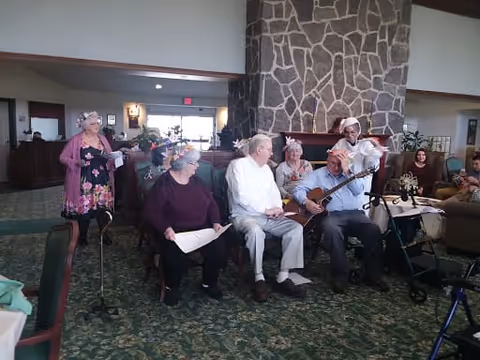A group of elderly people seated and standing in a common area with a large stone fireplace in the background. One person is playing a guitar while others hold papers, possibly singing or participating in a group activity. The room has carpeted floors and comfortable seating.
