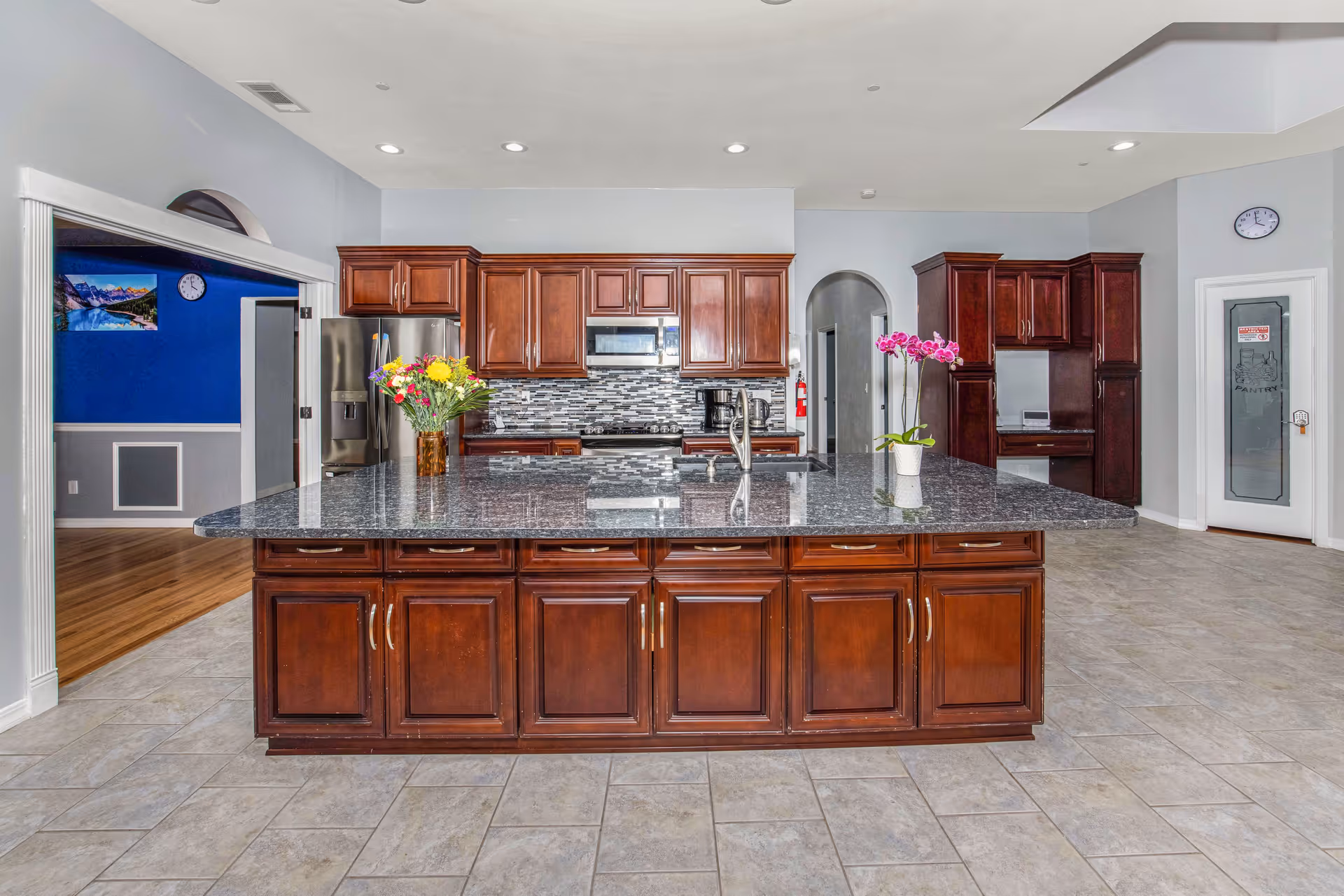 A spacious kitchen with a large granite island in the center featuring a sink and two flower vases. The kitchen has dark wooden cabinets, a stainless steel refrigerator, microwave, and coffee maker. The floor is tiled, and there is a pantry with a frosted glass door on the right. A blue-walled room with wooden flooring is visible through an open doorway on the left.