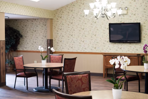 A dining area with round tables and chairs featuring red patterned upholstery. Each table has a white orchid plant in a white pot. The walls have floral wallpaper with a wooden chair rail. A flat-screen TV is mounted on the wall above a wooden sideboard. A chandelier with multiple lights hangs from the ceiling.