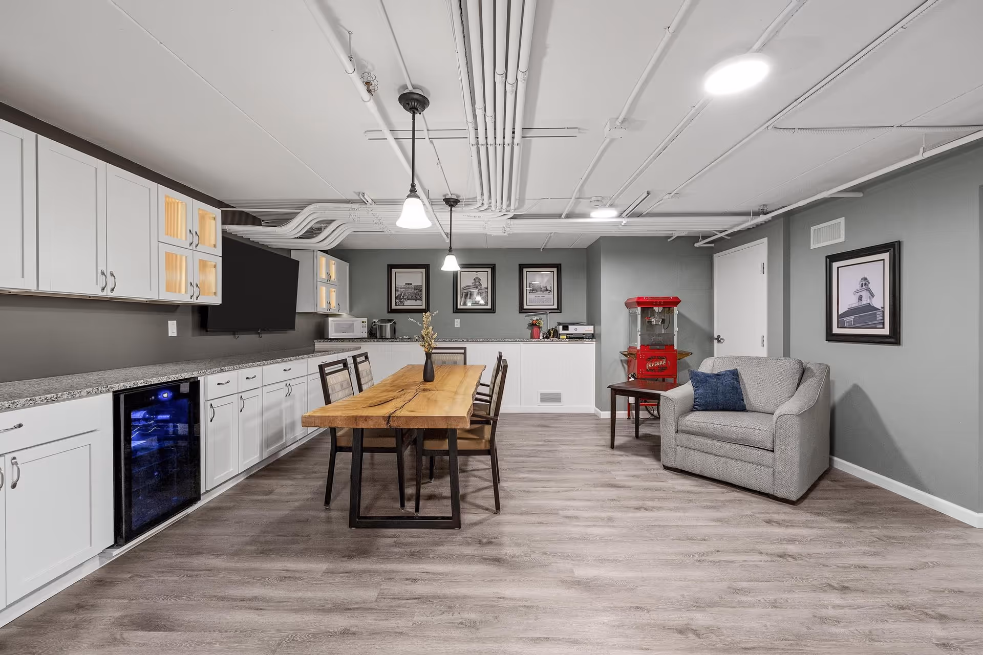 A modern common area with a long wooden table and chairs in the center, white cabinets with granite countertops along the left wall, a small wine cooler, a mounted flat-screen TV, and a gray armchair with a blue pillow on the right. The walls are painted gray and decorated with framed black and white photos. A red popcorn machine sits on a small table near the armchair. The ceiling has exposed white pipes and hanging light fixtures.