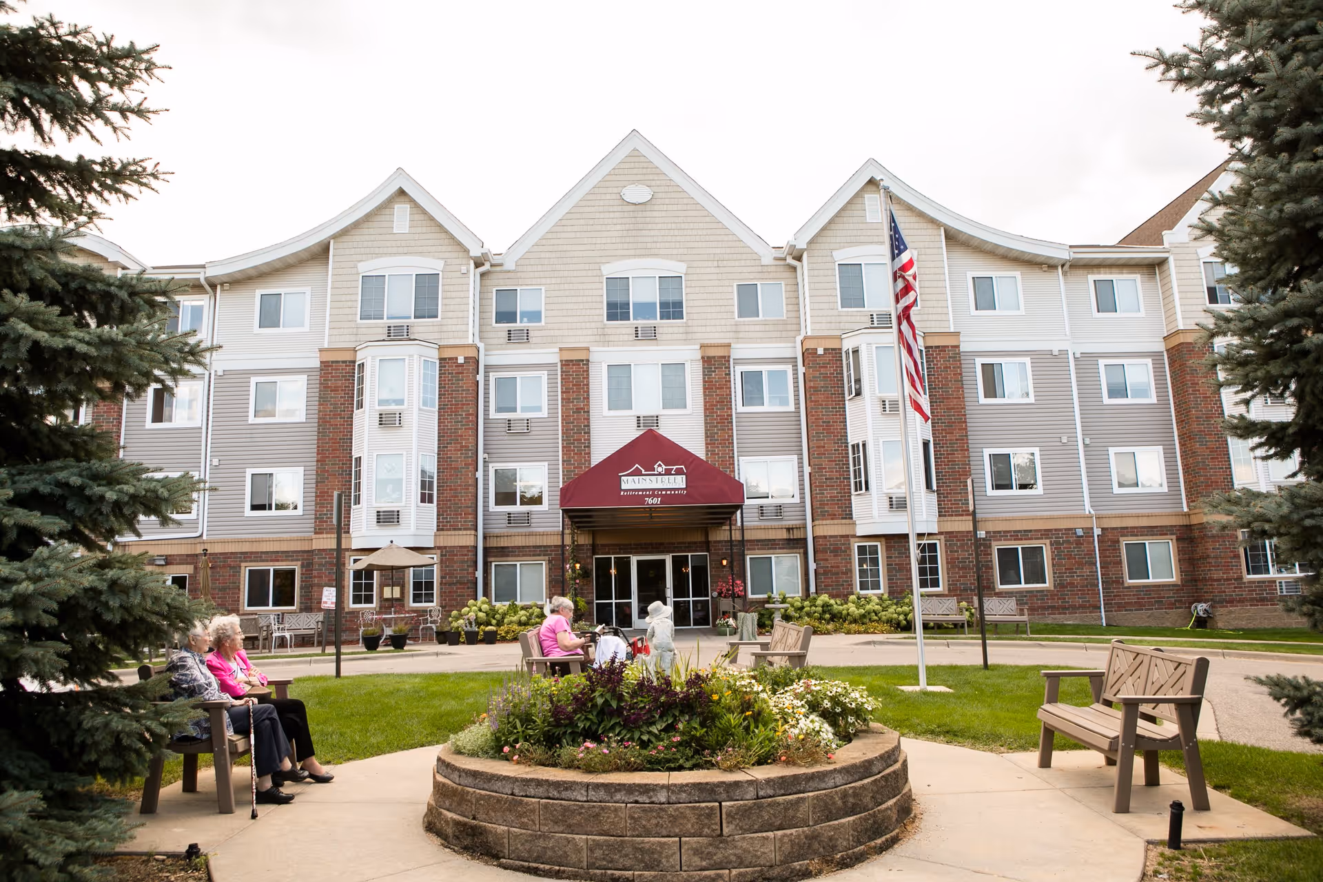Front exterior view of Mainstreet Village senior living facility with a circular flower bed in the center, benches around it, an American flag on a pole, and several elderly people sitting on benches in the outdoor area.