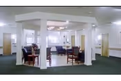 Communal lounge in a nursing facility with a central columned seating area, chairs, tables and windows.