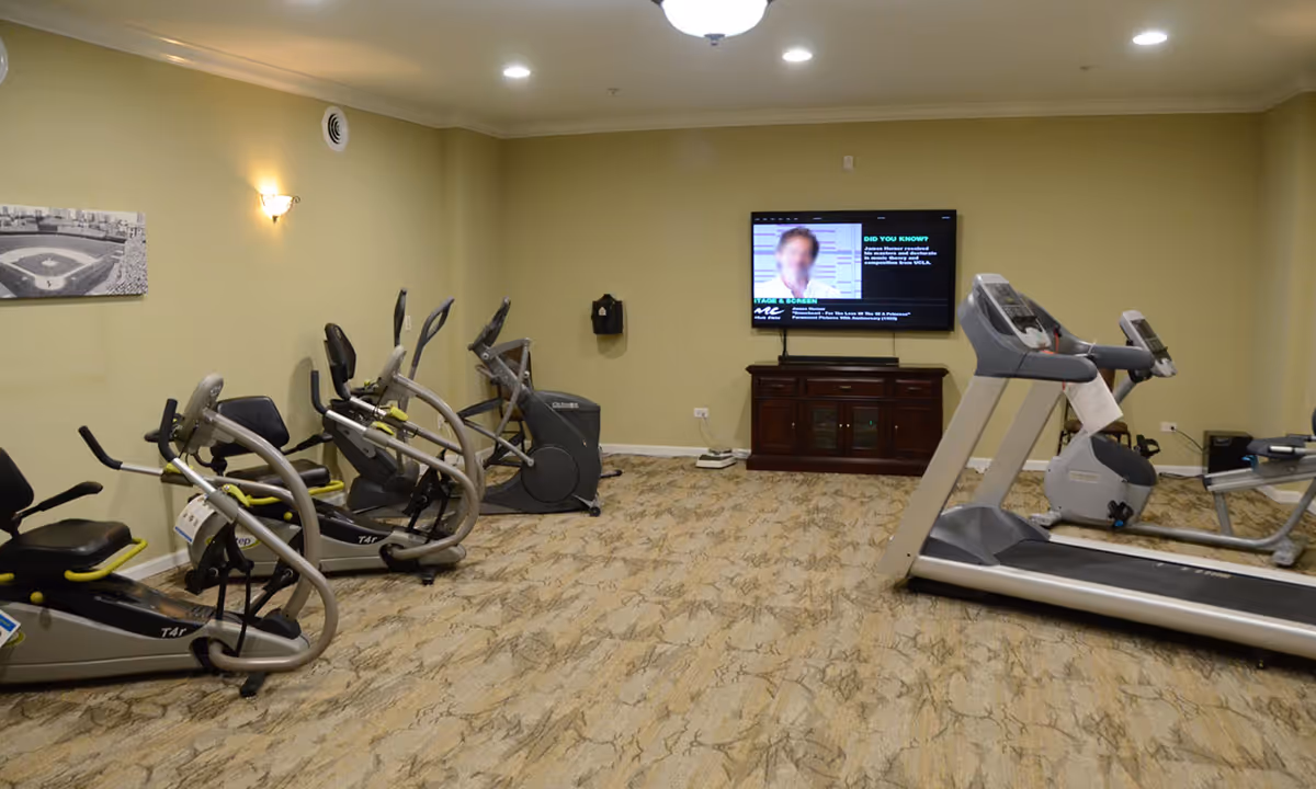 Exercise room with treadmills, stationary bikes, and a wall-mounted TV in a senior living facility.