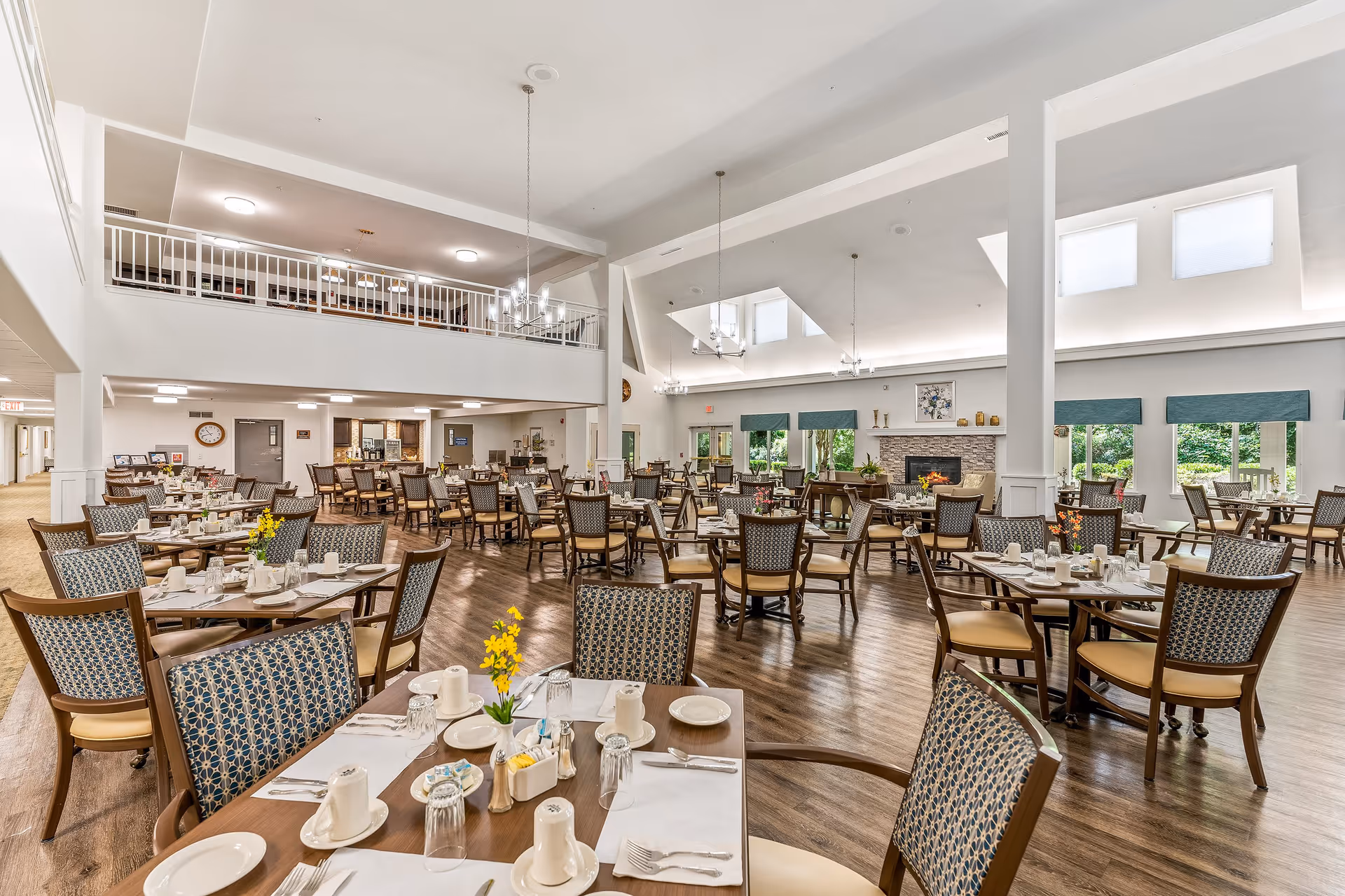 Spacious dining room with multiple round tables set with white placemats, cups, plates, and silverware. Chairs with patterned upholstery surround the tables. Large windows with green valances allow natural light to fill the room. A stone fireplace is visible on the far wall, and chandeliers hang from the high ceiling. The room has a warm wooden floor and an upper balcony area overlooking the dining space.