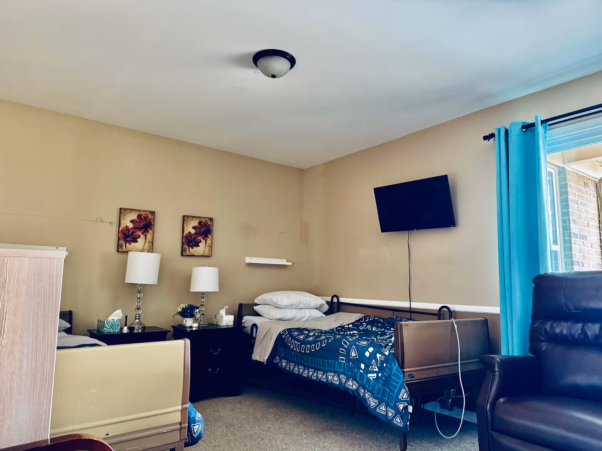 A bedroom in Aiello Adult Foster Care featuring two single beds with blue patterned blankets, two bedside tables with lamps and tissue boxes, a wall-mounted TV, a large window with blue curtains, and a dark brown recliner chair.