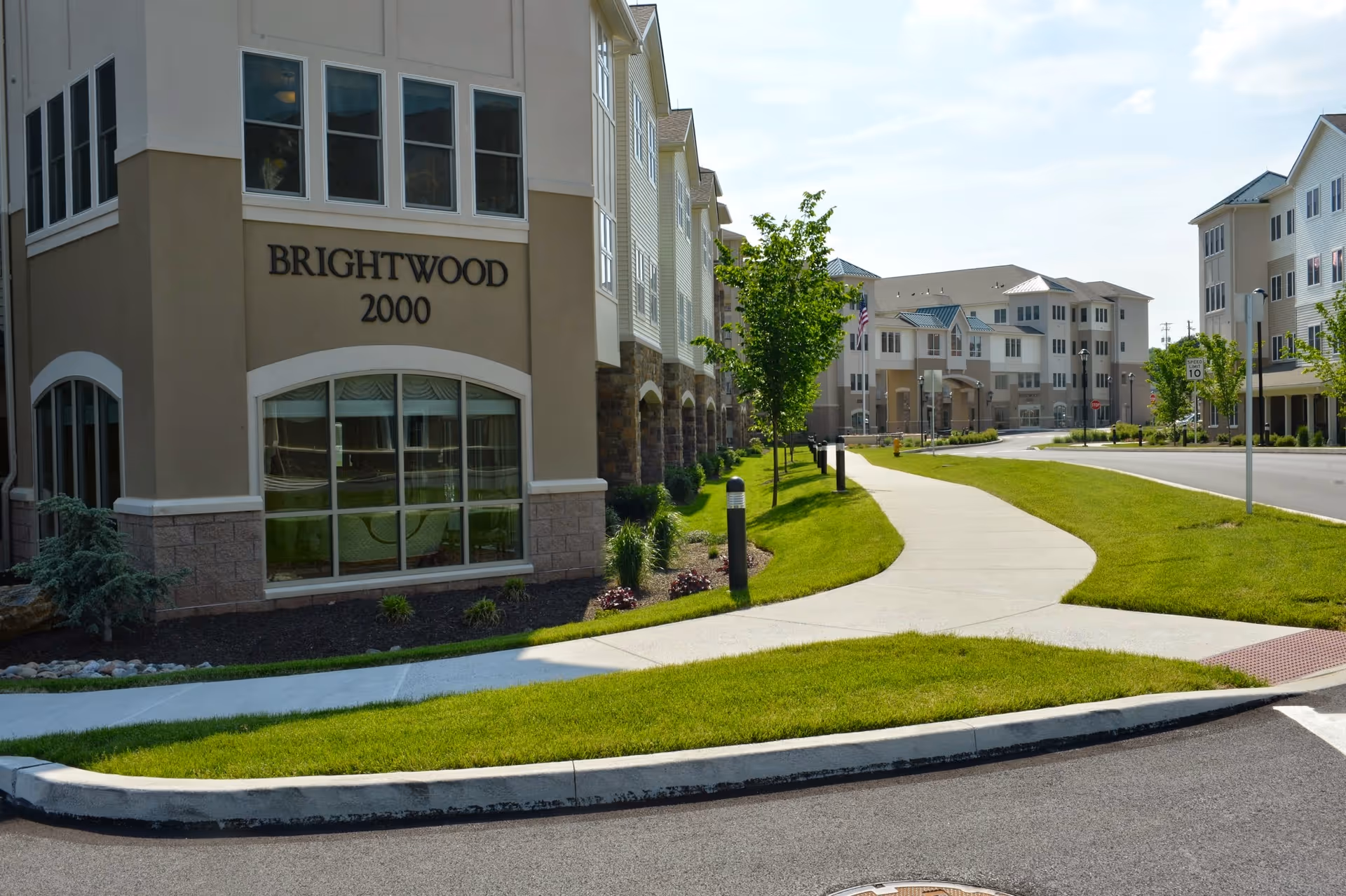 Exterior view of a senior living community named Brightwood 2000, showing multiple multi-story buildings with beige and stone facades, a curved sidewalk, green lawns, small trees, and a clear sky.