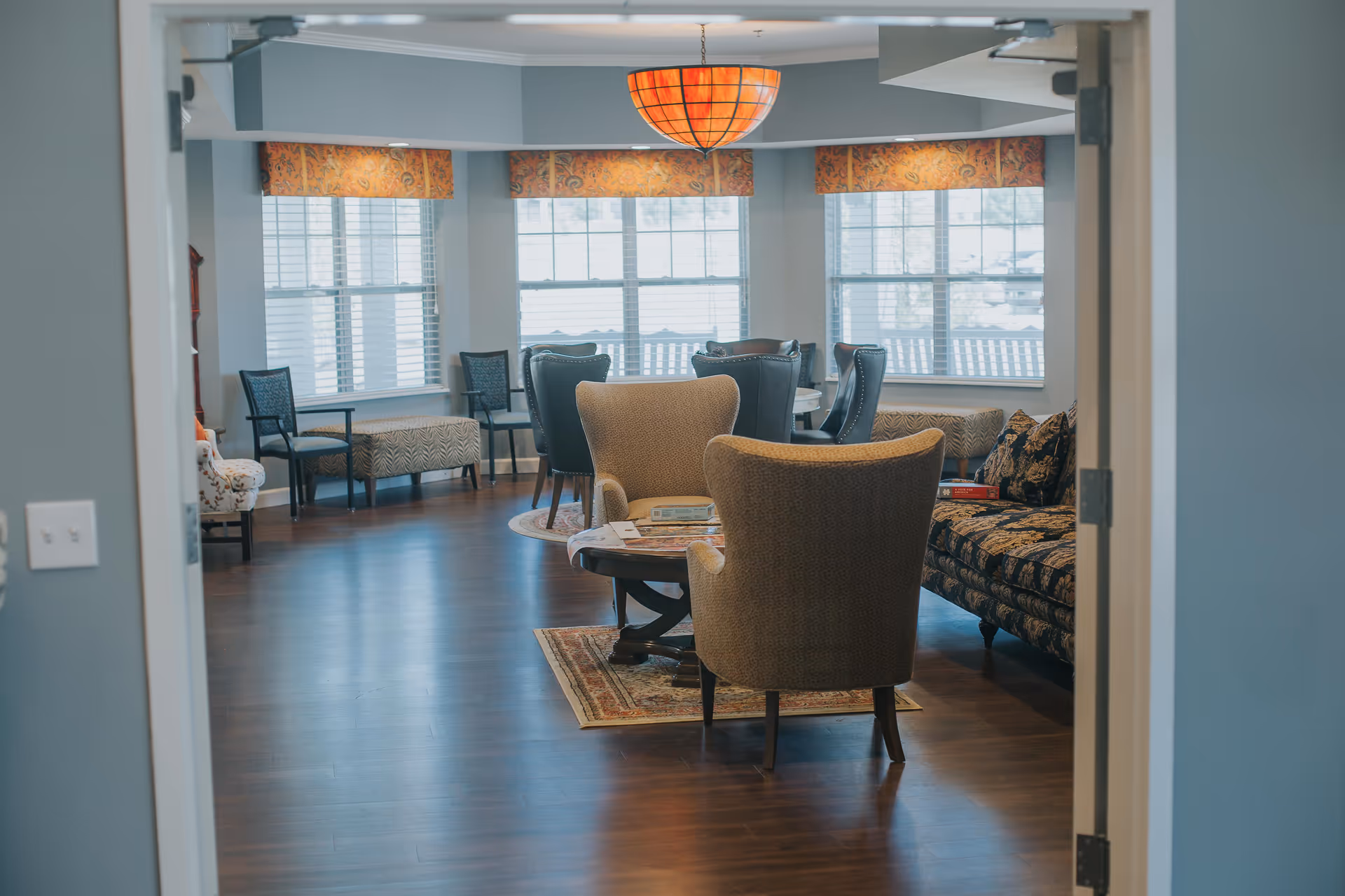 View through a doorway into a cozy living room area with two upholstered armchairs around a wooden coffee table on a small rug, a patterned sofa with cushions, and a dining table with chairs near large windows with blinds and orange valances. The room has wooden flooring and a warm hanging light fixture.