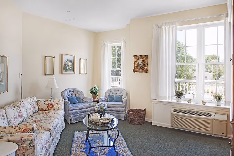 A cozy living room with a floral patterned sofa, two blue upholstered armchairs, a glass coffee table with a tea set, and a blue patterned rug. The room has cream-colored walls with framed artwork, two large windows with white curtains letting in natural light, and a basket near the window air conditioning unit.