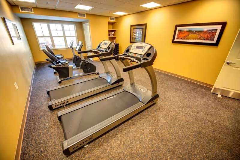 A small fitness room with two treadmills and two stationary bikes facing a window. The room has yellow walls, carpeted floor, and framed pictures on the walls. There is a door on the right side and a small shelf with weights in the corner.