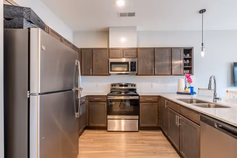 Modern kitchen with stainless steel refrigerator, oven, and microwave. The kitchen features dark wooden cabinets, a light-colored countertop with a double sink, and a pendant light hanging from the ceiling. The floor is wooden, and there are some decorative items including a pink flower on the counter.