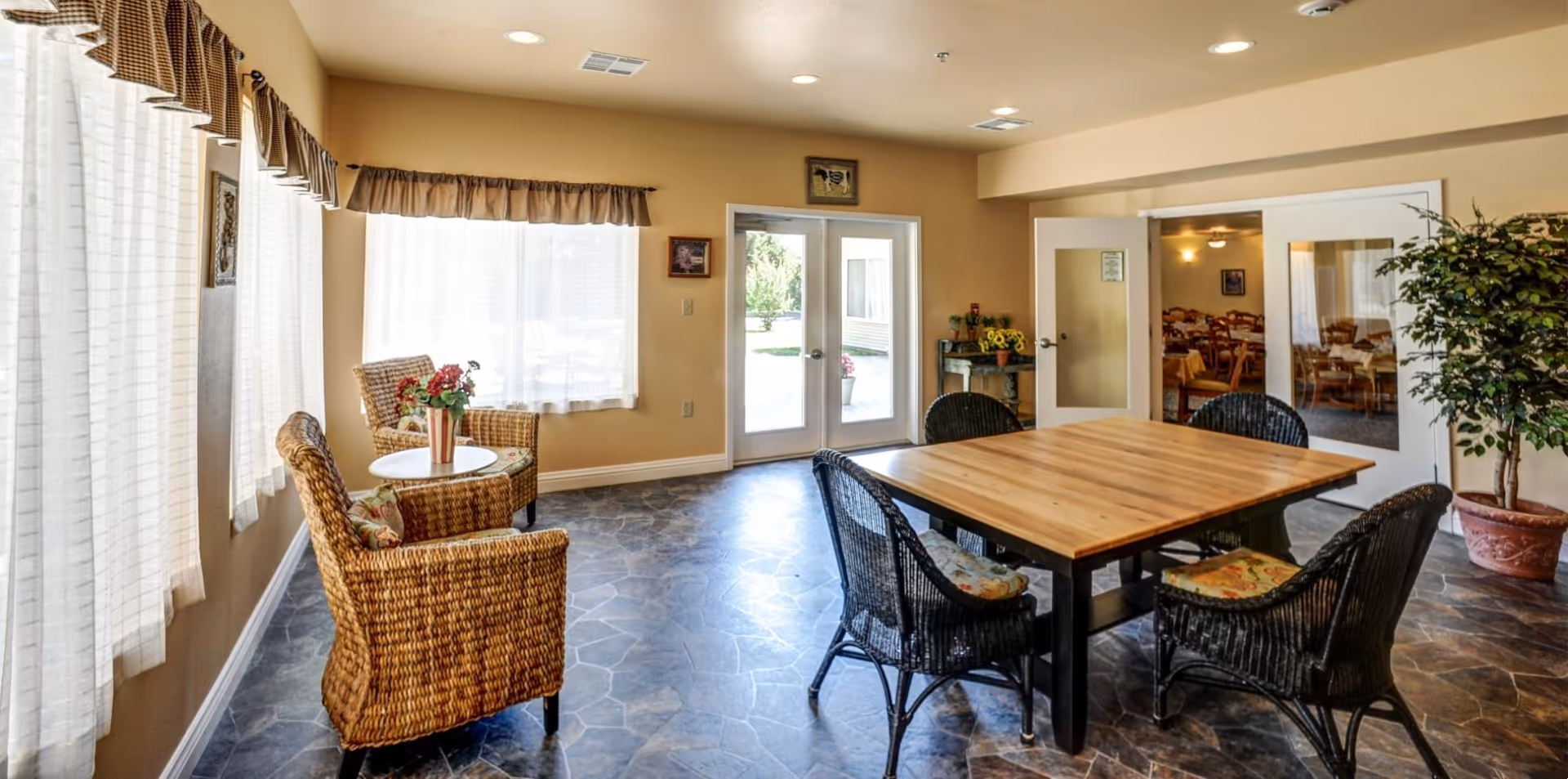 A bright room with large windows covered by sheer white curtains and brown valances. The room features a wooden table surrounded by four black wicker chairs with floral cushions. To the left, there are two wicker armchairs with floral cushions and a small round table holding a flower vase. Double glass doors lead outside, and a potted plant is placed near the right wall. Through an open door, another room with multiple dining tables and chairs is visible.