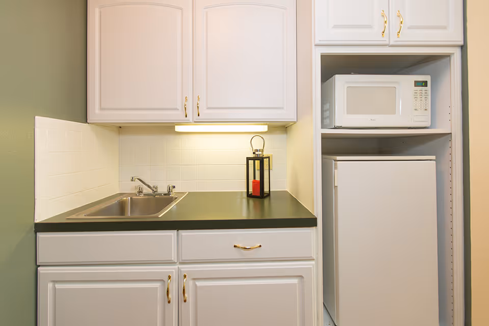 Small kitchenette area with white cabinets, a stainless steel sink, a black countertop, a white microwave on a shelf, and a white mini refrigerator below the microwave. There is a small decorative lantern with a red candle on the countertop.
